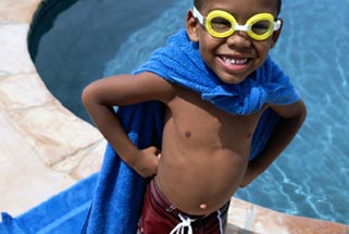 small black kid wearing a towel like a cape standing beside a pool with a big grin on his face.